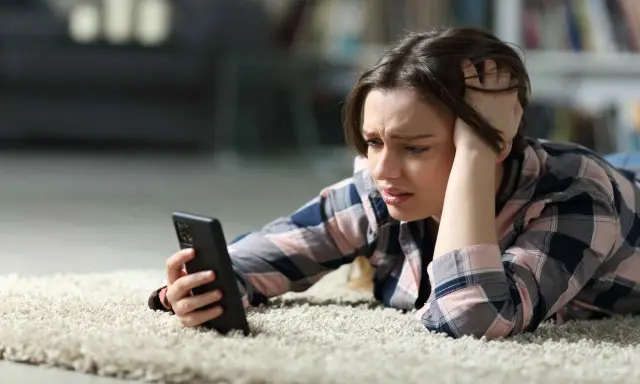 Zmartwiona dziewczyna leży na dywanie, próbując włączyć telefon, który nie chce się włączyć.
