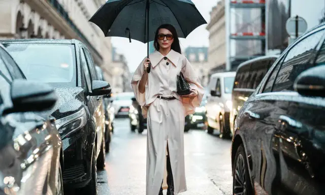 A woman in a chic cream outfit and sunglasses holds an umbrella, showcasing aesthetic rainy day outfits amidst a Parisian street scene with cars.