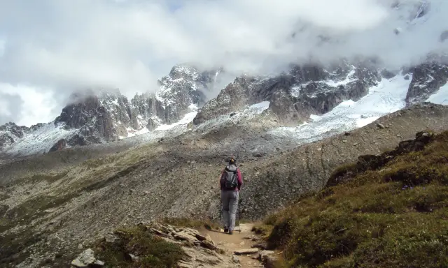 Osoba z plecakiem i kijkami idzie ścieżką w górach. W tle majestatyczne szczyty Alp, częściowo pokryte śniegiem i chmurami. Wspaniały widok na trekking w alpach.