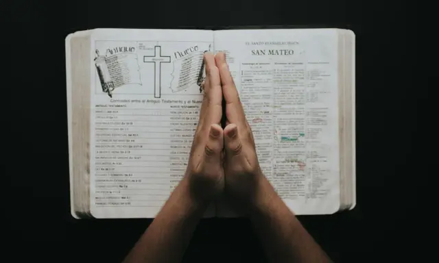 Dłonie złożone w modlitwie nad otwartą Biblią, z tekstami do modlitwy uwielbienia.