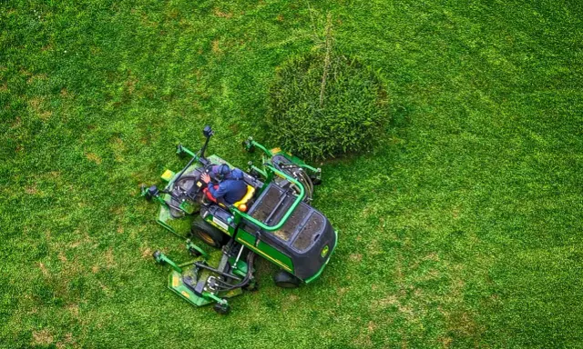 Un homme est assis sur un tracteur tondeuse, prêt à passer la tondeuse le dimanche. L'herbe est d'un vert éclatant.