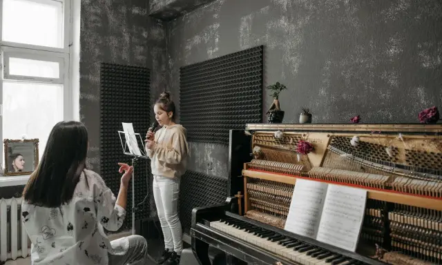 Dziewczynka ćwiczy swój **alt głos** przy mikrofonie, podczas gdy nauczycielka słucha jej uważnie. Obok stoi pianino z nutami.