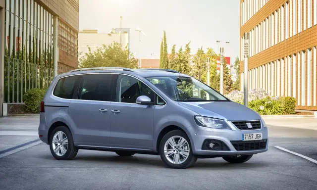 Srebrny Seat Alhambra, rodzinny van, parkuje przed nowoczesnym budynkiem z brązową elewacją.