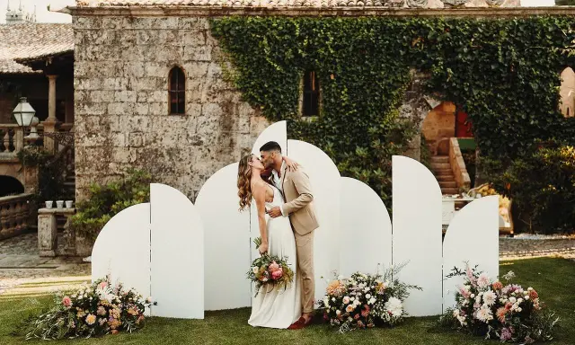 Novios besándose frente a un photocall moderno con arcos blancos y flores. ¡Ideas photocall boda únicas!