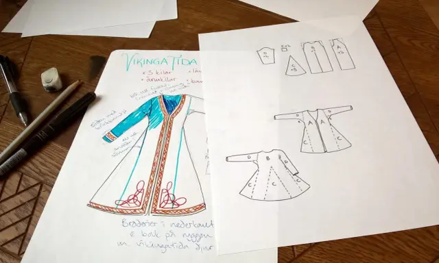 Skisser för en vikingatida kappa med mönster och sömnadsdetaljer. Pennor och suddgummi ligger bredvid.