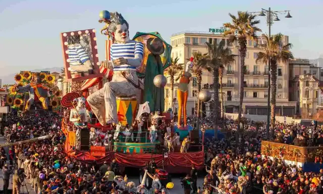 Carri allegorici e folla festante celebrano quando è Carnevale in Italia. Un clown gigante domina la scena, circondato da figure colorate e palme.