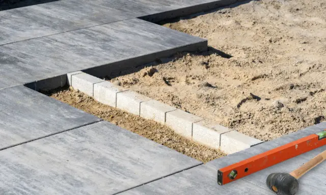 Budowa tarasu z płyt betonowych w toku. Widać układane płyty, podsypkę piaskową i obrzeża. Narzędzia budowlane leżą obok.