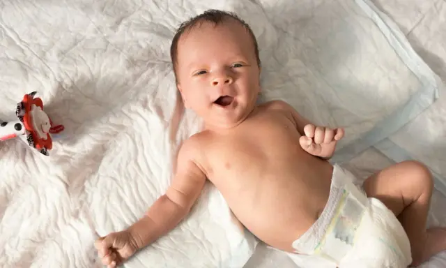 Baby mit offenem Mund, vielleicht im 6-Wochen-Schub, liegt auf einer weißen Unterlage neben einem bunten Spielzeug.