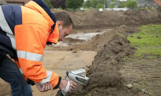 Pracownik w pomarańczowej kurtce wykonuje badanie gruntu za pomocą specjalistycznego urządzenia.