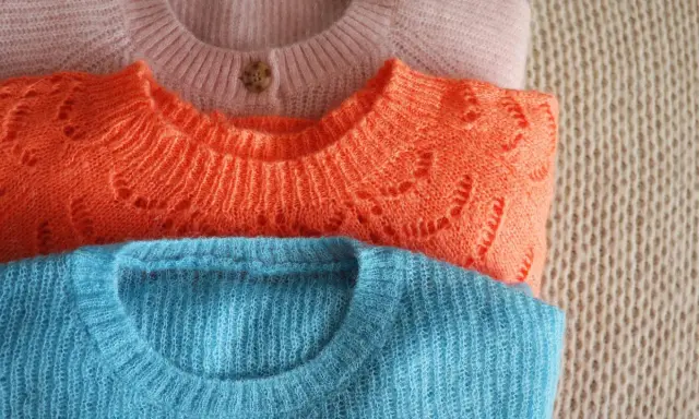 Drei gestapelte Pullover in Rosa, Orange und Blau. So pflegt man einen wollpullover richtig, damit er lange schön bleibt.