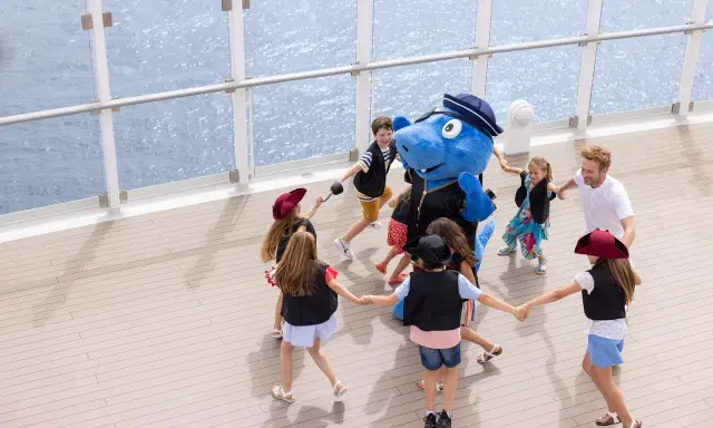 Bambini giocano in cerchio con un personaggio blu su una nave, con la costa toscana sullo sfondo.