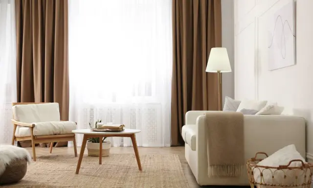 Salon cosy avec 1 ou 2 rideaux par fenêtre, fauteuil en bois, canapé blanc, table basse et tapis en jute.