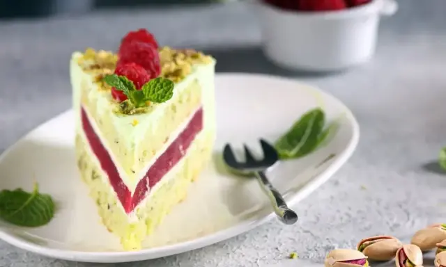 Pyszny tort pistacjowo malinowy z malinami i miętą na talerzu. Obok widoczne łuskane pistacje.