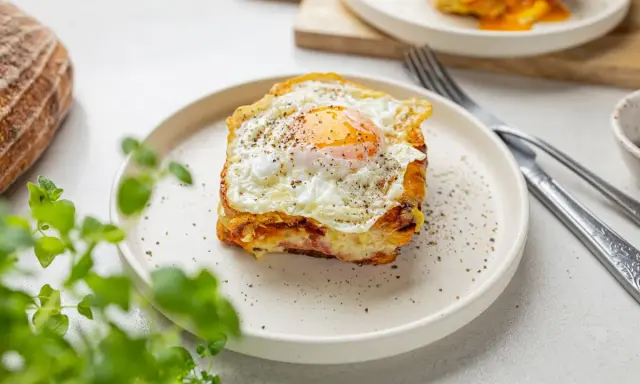 Pyszny croque madame z jajkiem sadzonym na wierzchu, na talerzu, obok chleba i zielonych liści.