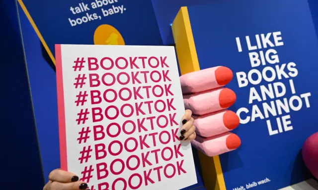 Mani che tengono un cartello con #BOOKTOK ripetuto. Sullo sfondo, un libro blu con la scritta "I LIKE BIG BOOKS AND I CANNOT LIE".
