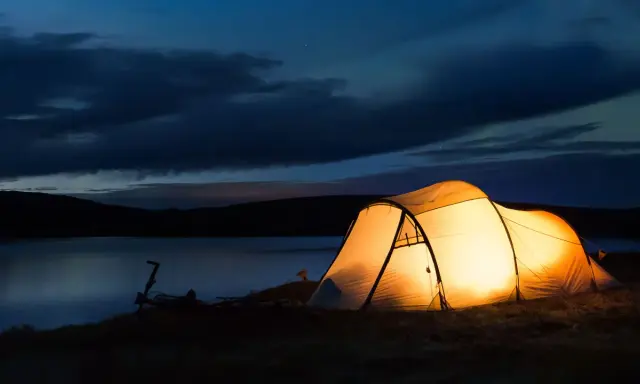 Upplyst tält vid en sjö i skymningen. En cykel står parkerad bredvid. Perfekt för camping Sverige karta-äventyr.