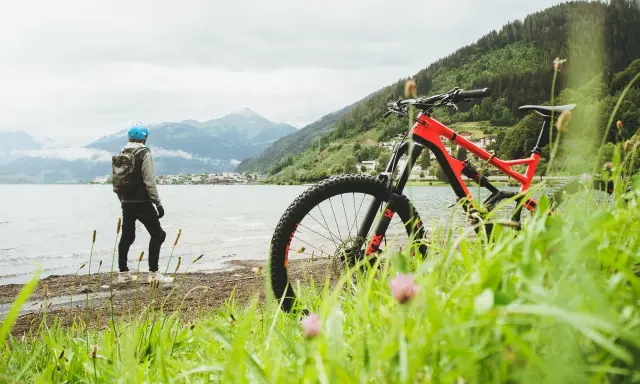 En cyklist med hjälm och ryggsäck står vid en sjö med berg i bakgrunden. En mountainbike står redo för äventyr på mtb leder.