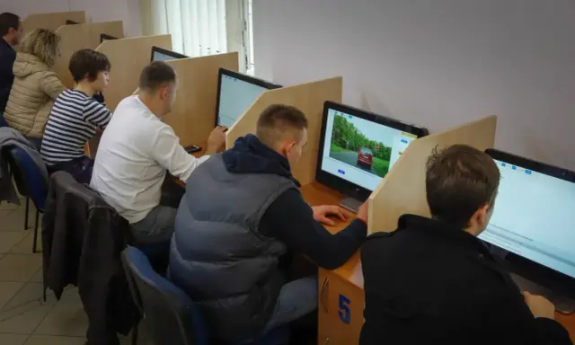 Ludzie siedzą przy komputerach, rozwiązując testy. Zastanawiają się, ile trzeba mieć punktów, żeby zdać teorię na prawo jazdy.