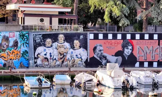 Graffiti colorati sul muro del borgo San Giuliano: astronauti con Alf, personaggi di Amarcord e cani. Barche ormeggiate sull'acqua riflettono i colori.