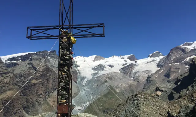 Croce metallica e di pietra in cima a una montagna, con un ghiacciaio e vette innevate sullo sfondo. Il **palon di Resy** domina il panorama.