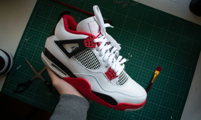 Białe buty Jordan 4 z czerwonymi akcentami, które wyglądają jak podróbki. Na stole leżą nożyczki i pędzel.