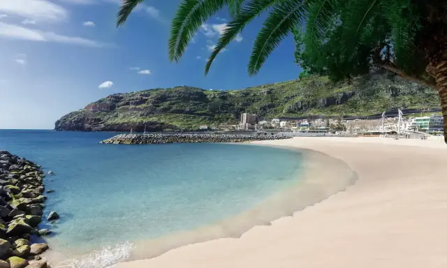 Traumhafter Madeira Strand mit türkisfarbenem Wasser, goldenem Sand und grünen Hügeln im Hintergrund. Palmen spenden Schatten.
