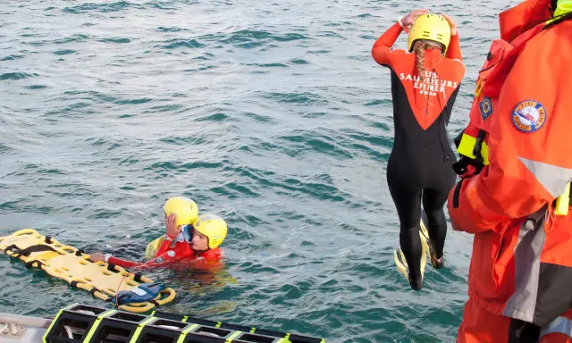 Sauvetage en mer : une sauveteuse en combinaison noire et orange se prépare à plonger. Deux personnes en gilets de sauvetage et casques jaunes flottent près d'une civière. Communication VHF et MMSI essentielle.