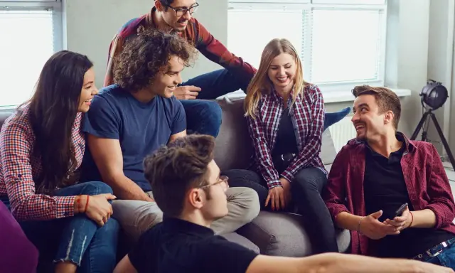 Grupa przyjaciół śmieje się, siedząc na kanapie. Wyglądają, jakby grali w grę, może nawet w Familiadę, odpowiadając na zabawne pytania.