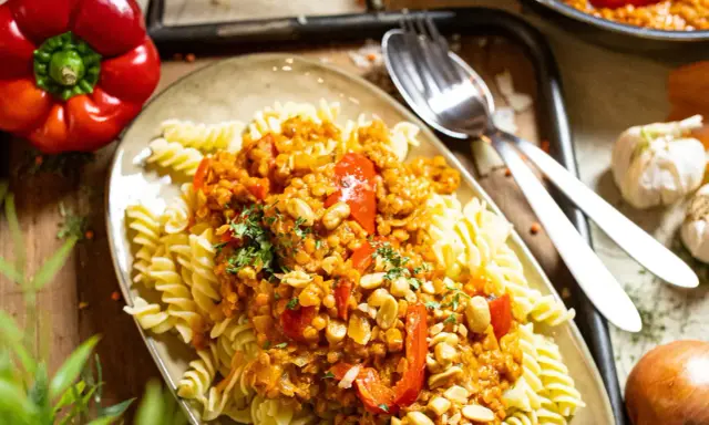 Fusilli mit einer herzhaften bolognese mit roten linsen, garniert mit Erdnüssen und Petersilie. Daneben liegen eine rote Paprika und Knoblauch.