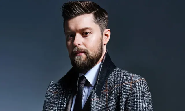 Mężczyzna z brodą i starannie ułożonymi włosami, w eleganckim płaszczu w kratę. Remigiusz Mróz wiek nie jest widoczny, ale styl sugeruje dojrzałość.
