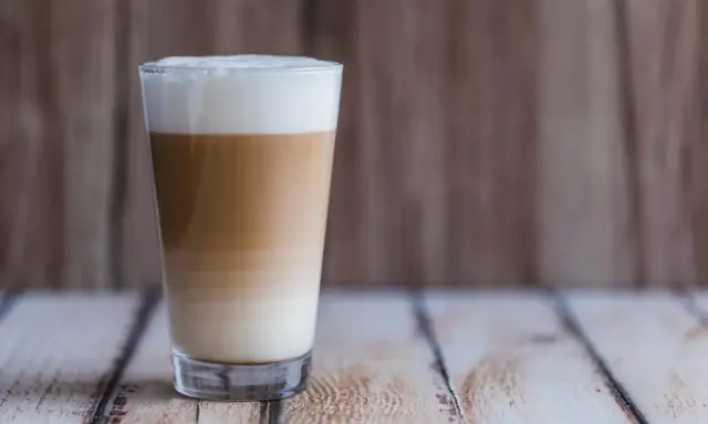 Ein hoher Latte Macchiato mit Mandelmilch, geschichtet in drei Farben, steht auf einem Holztisch.