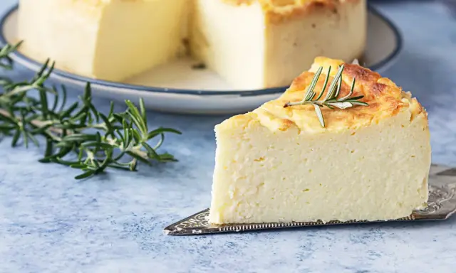 Una fetta di torta di ricotta soffice e dorata, guarnita con rosmarino, su un piatto da torta argentato.