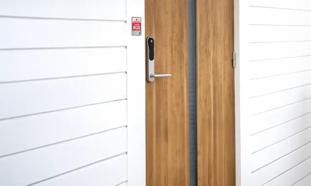 En modern ytterdörr med ett Verisure Yale Doorman V2N lås. Dörren är i trä och har en vitmålad panelvägg bredvid.