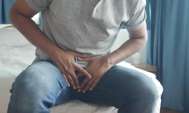 Mężczyzna siedzi na łóżku, trzymając się za podbrzusze. Szuka informacji, jak naturalnie zmniejszyć prostatę.