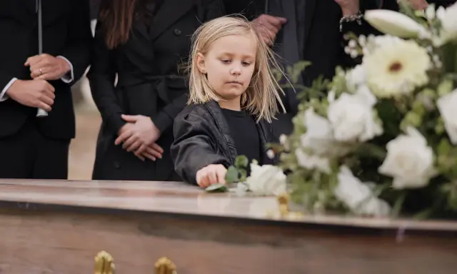 Smutne dziecko na pogrzebie, dotyka kwiatów na trumnie. W tle dorośli w czerni. Czy to dziecko wierzy w przesądy?