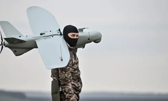 Mann in Tarnkleidung und schwarzer Sturmhaube trägt eine Drohne. Seit wann gibt es Drohnen? Die Geschichte reicht weit zurück.