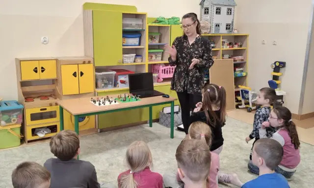 Nauczycielka prowadzi zajęcia kreatywne dla dzieci. Na stole przed nimi leżą klocki Lego, a dzieci z uwagą słuchają.