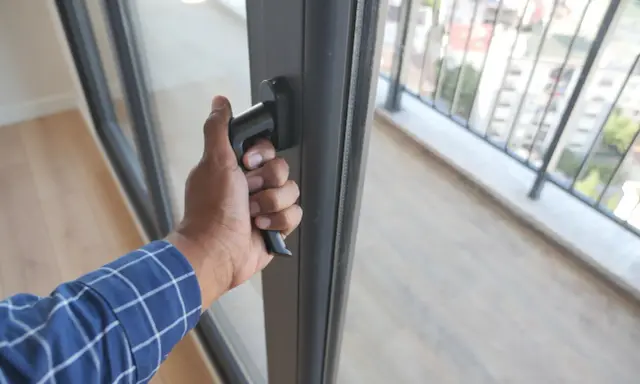 Dłoń chwyta za klamkę, wykonując regulację drzwi balkonowych dwuskrzydłowych. Widok na balkon i miasto.