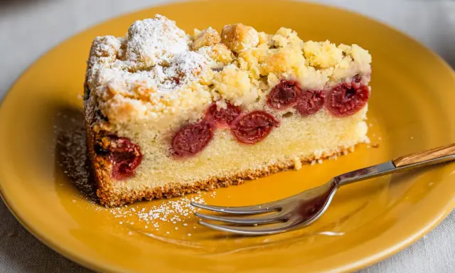 Ein saftiger Kirschkuchen aus der Springform, bestäubt mit Puderzucker und Streuseln, auf einem gelben Teller mit einer Gabel.