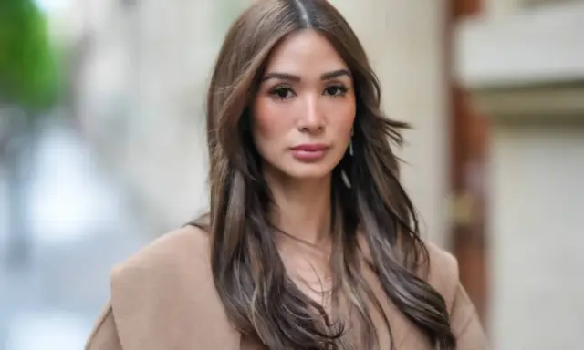 Mujer con cabello castaño largo y ondulado, viste un abrigo beige. Su maquillaje es sutil, un estilo "old money" que realza su belleza natural.
