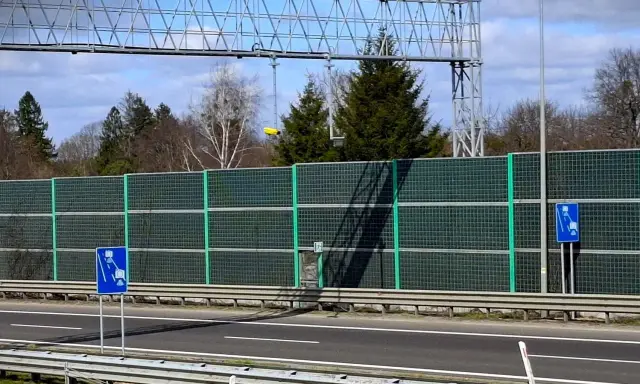 Znak drogowy informujący o odcinkowym pomiarze prędkości na tle ekranów akustycznych i drzew.