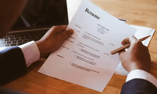 Dłoń trzymająca długopis nad dokumentem CV obok laptopa.
