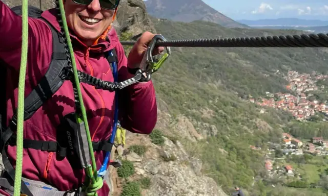 Donna sorridente si aggrappa a un cavo d'acciaio, godendosi un'avventura sulle ferrate Val di Susa con un panorama mozzafiato.