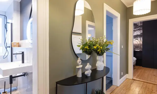 Élégant couloir maison avec miroir asymétrique, vases décoratifs, bouquet de fleurs jaunes et vue sur une salle de bain et une chambre.