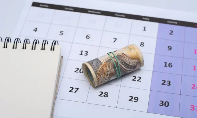 Zrolowane banknoty na kalendarzu, symbolizujące ekwiwalent czasu i pieniędzy. Obok pusty notatnik.
