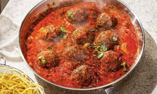 Frikadeller i tomatsås som puttrar i en panna, med en sidokorg med spaghetti.