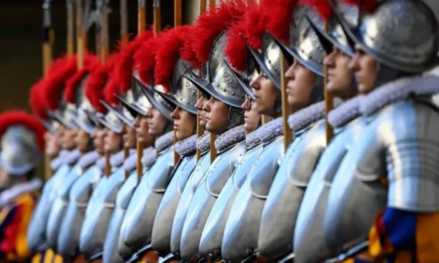 Guardie svizzere in fila, con elmi e piume rosse. Curiosità sulla loro uniforme storica e il loro ruolo.