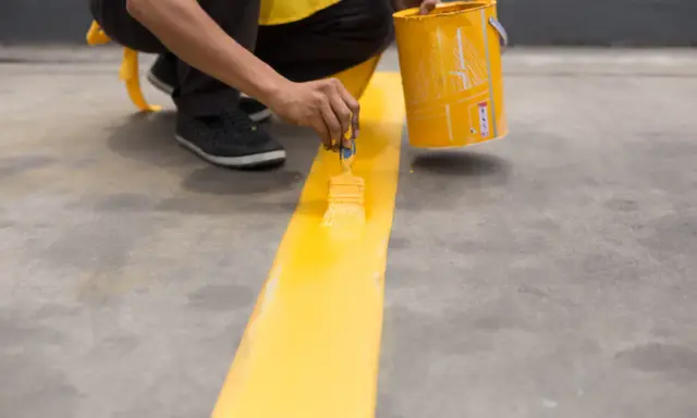 Persona pintando una línea amarilla sobre suelo de cemento sin pulir, usando una brocha y un cubo de pintura.