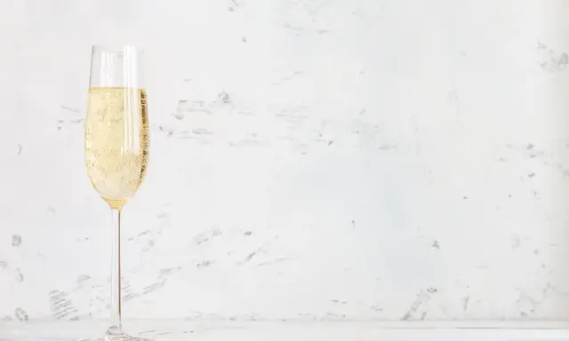 Ett glas champagne med bubblor, kanske en Jaques Selosse, står på ett vitt bord mot en ljus, texturerad bakgrund.