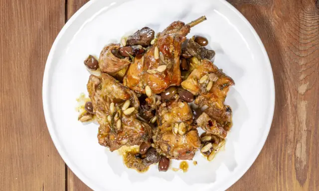 Pezzi di coniglio alla ligure, cotti con olive, pinoli e erbe aromatiche, serviti su un piatto bianco.